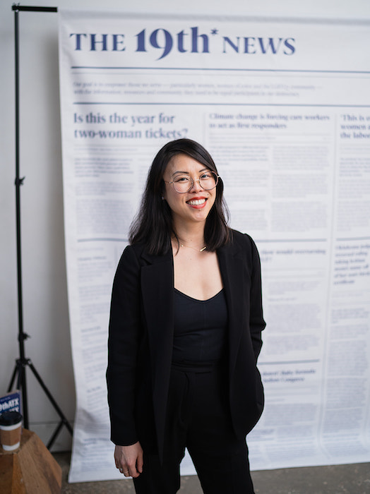 Julia B. Chan is pictured in black pants and a black blazer standing in front of a newspaper print backdrop that says The 19th News. Photo by Lauren Slusher for The 19th 